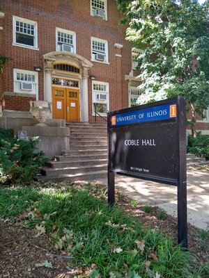 A historic brick building with shallow concrete steps leading up to an ornate door. A sign in the foreground says "Coble Hall."